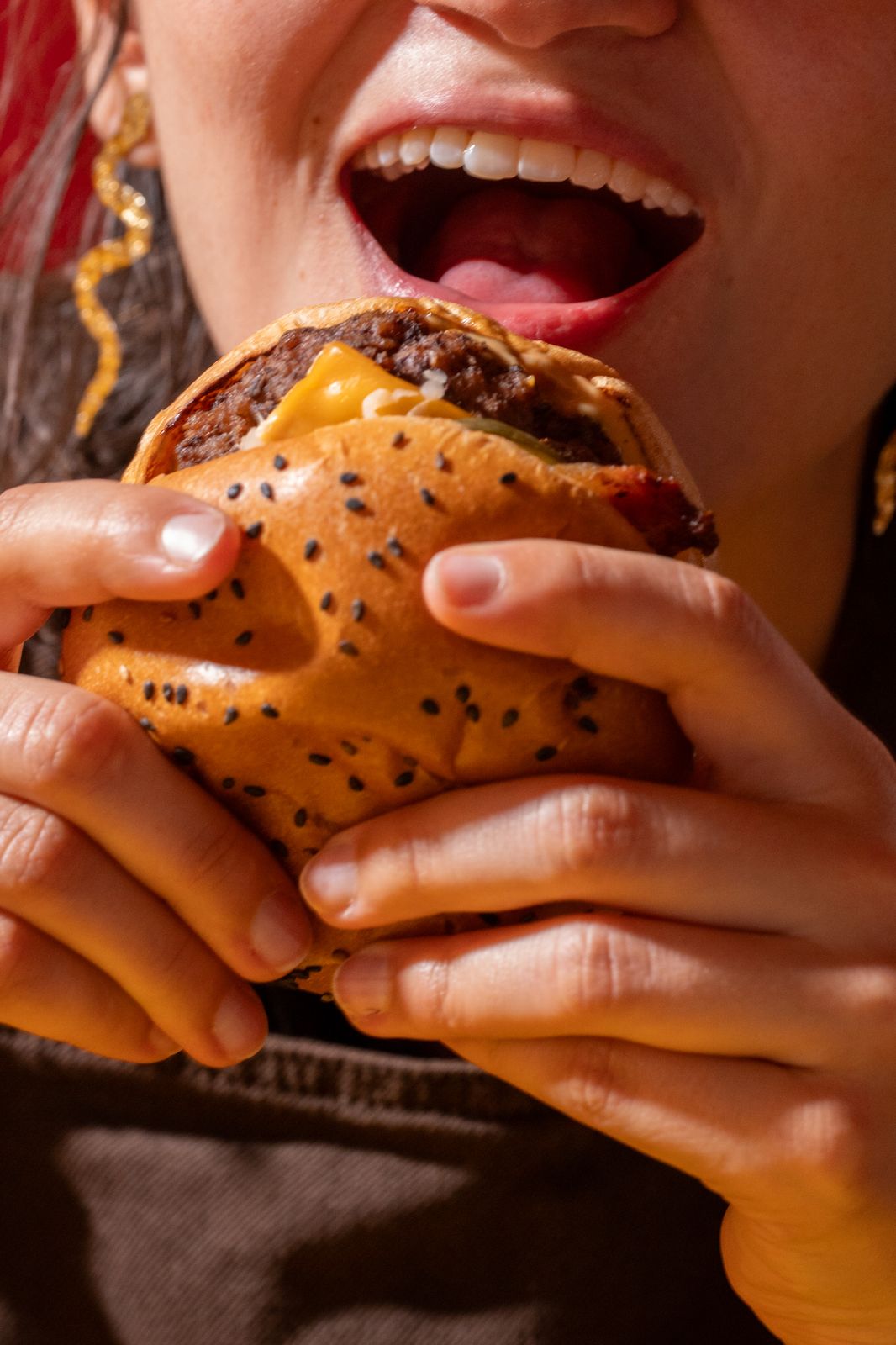 A person holding a sesame bun burger with both hands, mouth open for a bite