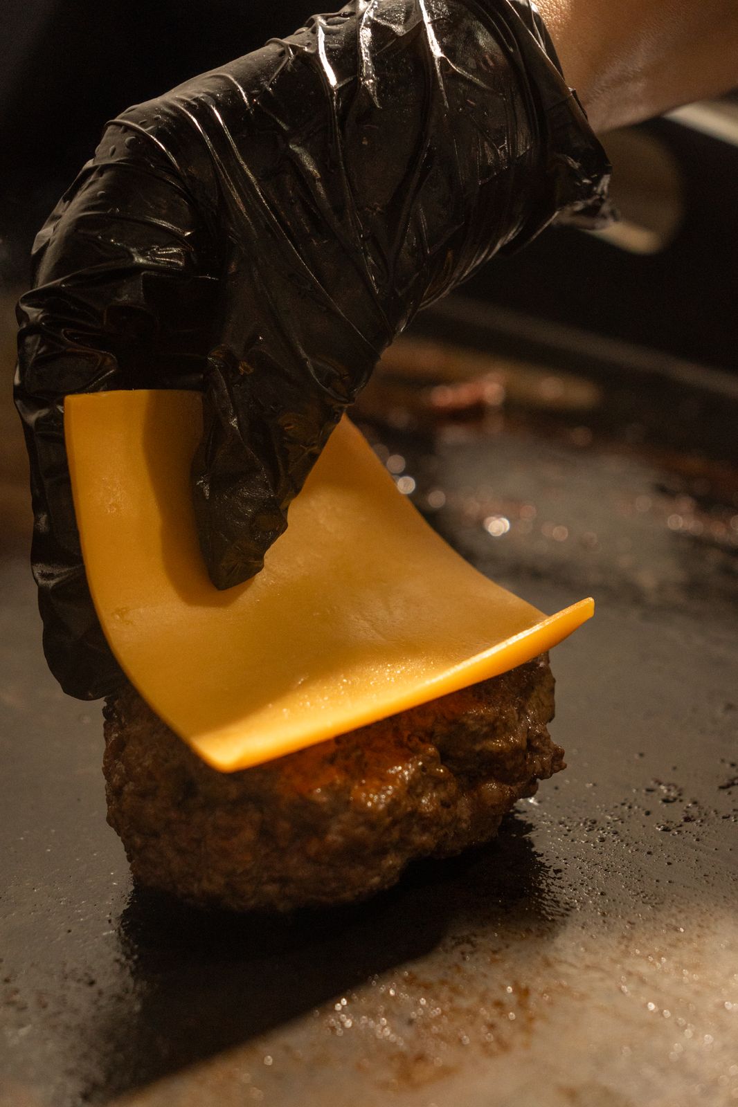 A gloved hand laying a slice of American cheese on a patty on the grill